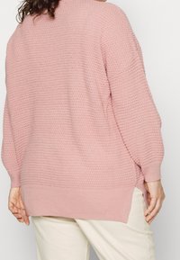 Jasnoróżowy, oversize sweter z teksturowanym wzorem dzianiny, bocznymi rozcięciami i luźnym krojem. Prążkowane mankiety i dół dodają wyjątkowego akcentu.