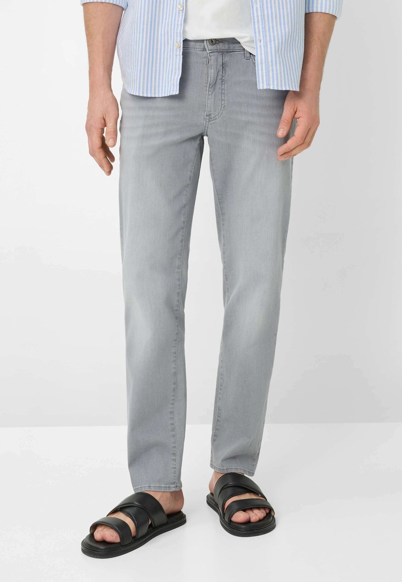 Man draagt lichtgrijze jeans met rechte pijpen, zwarte sandalen met dubbele bandjes, wit T-shirt en blauw gestreept overhemd met knopen.