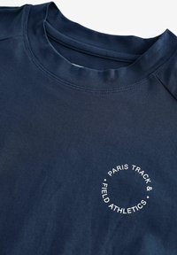 Cămașă sport de culoare albastru marin, cu o textură netedă, având un imprimeu rotund alb care spune "PARIS TRACK & FIELD ATHLETICS" pe față.
