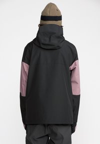 Veste rectangulaire noire et rose avec capuche, présentant une texture mate, des manches contrastées et une coupe décontractée. Un bonnet beige est porté.