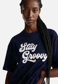 Námořnická modrá bavlněná trička s krátkým rukávem s nápisem "Stay Groovy" v tučném bílém písmu. Měkká textura a uvolněný střih.