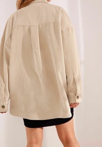 Beigenvärinen, kohoraitainen, ylivuotava paito, jossa on suuri kaulus ja napitettu hiha, puettu mustan vaatteen päälle.