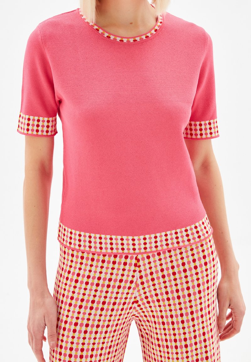 Rosa kortärmad topp med texturerad väv, med en kontrasterande dekorativ kant av små röda och gula mönster vid halsringningen och ärmarna.