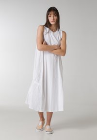 Vestido de algodón blanco sin mangas con un corte relajado, que presenta un detalle fruncido en un lado y un dobladillo fluido, combinado con zapatillas de color claro.