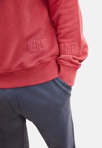 Rode sweatshirt met reliëf tekst "ELB" en "IND" op de mouwen, gecombineerd met marineblauwe sweatpants, beide met een gladde, gestructureerde stof.