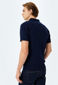 Mörkblå stickad polo med ribbad textur, korta  ärmar och figursydd design. Bärs med mörkblå jeans. Minimalistisk stil.