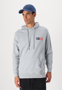 Grå hættetrøje med frontlomme, indeholdende et flerfarvet logo-patch. Sammen med en blå baseballkasket. Glat tekstur.