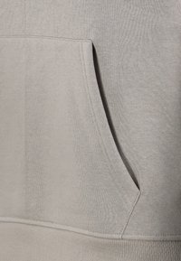 Hellgrauer Pullover aus strukturiertem Stoff, mit einer Kängurutasche und geripptem Saum. Einfaches Design mit minimalen Nähten.