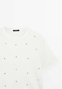 T-shirt blanc à manches courtes avec de petits œillets métalliques argentés disposés uniformément sur le devant et un col rond.