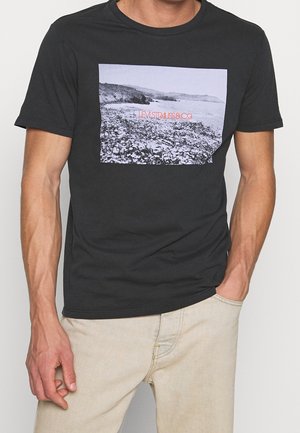 Mand iført en sort T-shirt med et sort-hvidt kystlandskab og orange "Levi Strauss & Co." tekst foran.