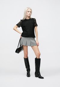 Μαύρο δαντελένιο τοπ με κοντά μανίκια, γκρι πλισέ φούστα, μαύρες μπότες μέχρι το γόνατο και μικρή μαύρη τσάντα χειρός· μινιμαλιστικός σχεδιασμός και ομαλές υφές.