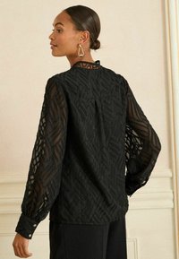 Bluză neagră texturată cu un model în valuri, mâneci lungi semi-transparente și manșete cu nasturi. Prezintă o închidere la gât în spate. Design simplu.