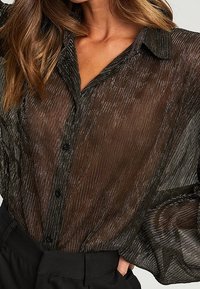 Blusa nera trasparente con un motivo a righe testurizzato, chiusura a bottoni e colletto a punta. Maniche lunghe con dettagli arricciati.