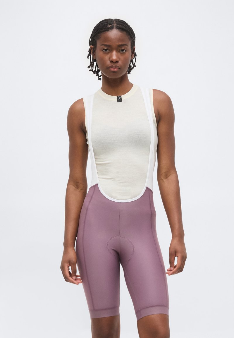 Νεαρή γυναίκα με κοντά πλεγμένα μαλλιά, που φοράει λευκό αμάνικο τοπ και μωβ ποδηλατικά σορτς με τιράντες, στέκεται μπροστά σε απλό φόντο.