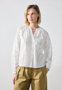 White button-up shirt featuring intricate embroidery on puff sleeves, with a smooth texture and slight sheen, paired with khaki pants.