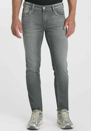 Uomo con jeans grigi slim-fit e sneakers bianche e grigie, in piedi contro uno sfondo semplice.