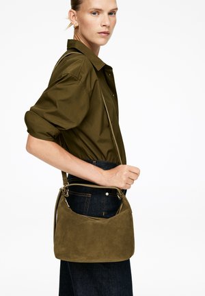 Olivgrünes Wildleder-Crossbody-Tasche mit geschwungenem Design, Reißverschluss und einem dünnen, verstellbaren Träger, getragen zu einem dunklen Outfit.