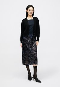 Cardigã preto sobre uma blusa azul-marinho, combinado com uma saia de lantejoulas azul-marinho transparente. A modelo usa sapatos de salto pretos e meias escuras. Um colar simples realça o visual.