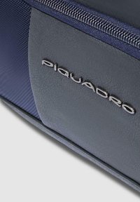 Bolso azul marino con material sintético texturizado, que presenta un acento de cuero suave y el logo plateado "Piquadro" cerca del zipper.