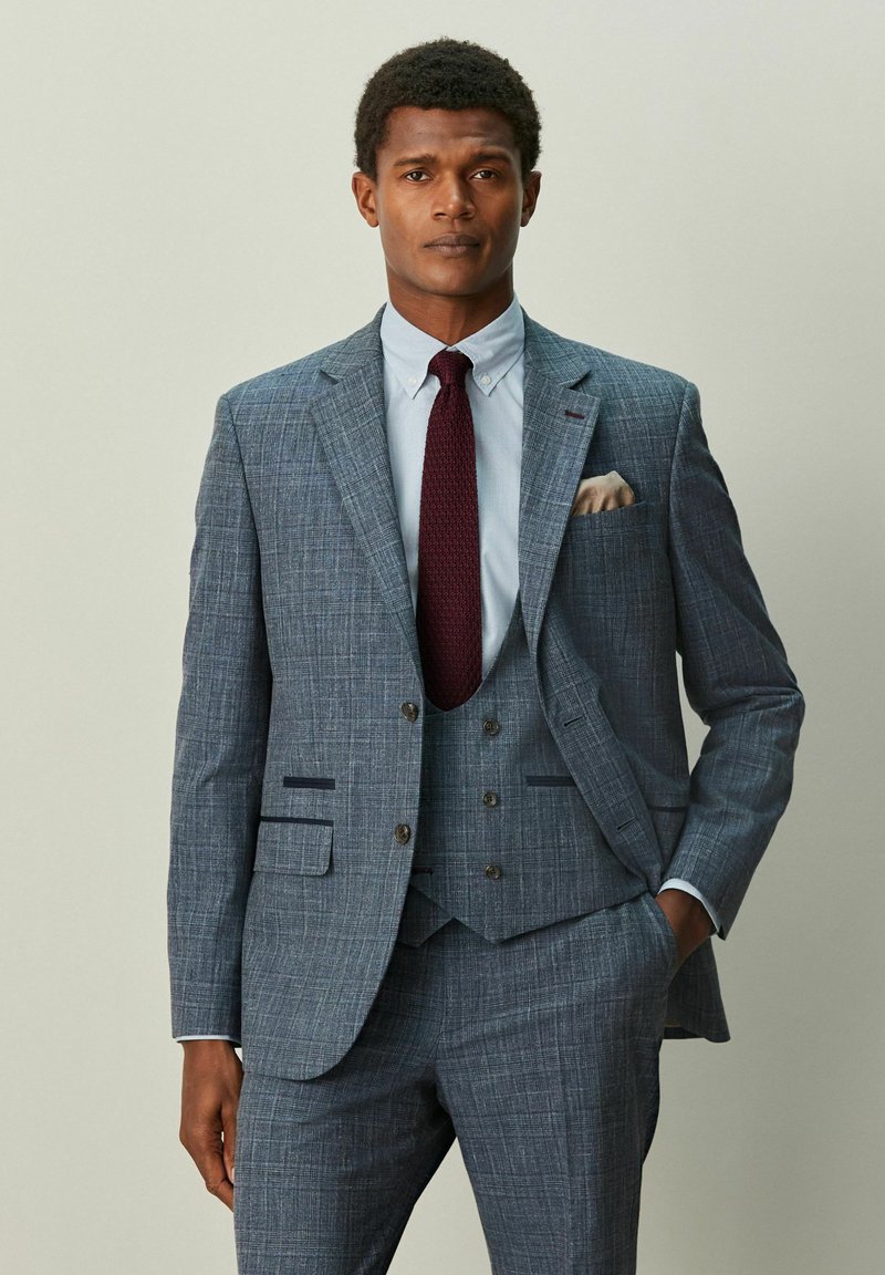 Grauer karierten Dreiteiler mit einem Stegkragen, einer Brusttasche mit einem hellfarbenen Taschentuch und einer bordeauxroten Strickkrawatte.