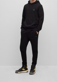 Zwarte hoodie met een voorzak en logo patch, gecombineerd met zwarte joggers met een logo, aangevuld met hoge sneakers met gele accenten.