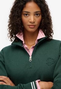 Donkergroene ritssluiting pullover van een gestructureerde stof, met daaronder een roestvrijstalen shirt in het roze. Accenten omvatten een logo en gestreepte manchetten.