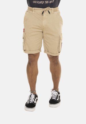 Homme portant un short cargo beige avec les ourlets retroussés, des baskets noires et blanches, et un t-shirt noir avec le texte "UNLEASHED THE WOLF."