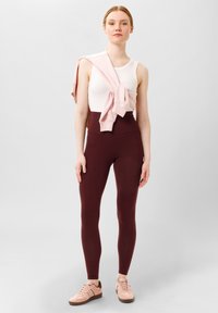 Débardeur blanc, leggings bordeaux taille haute, et hoodie zippé rose drapé sur les épaules ; associé à des baskets rose pâle.