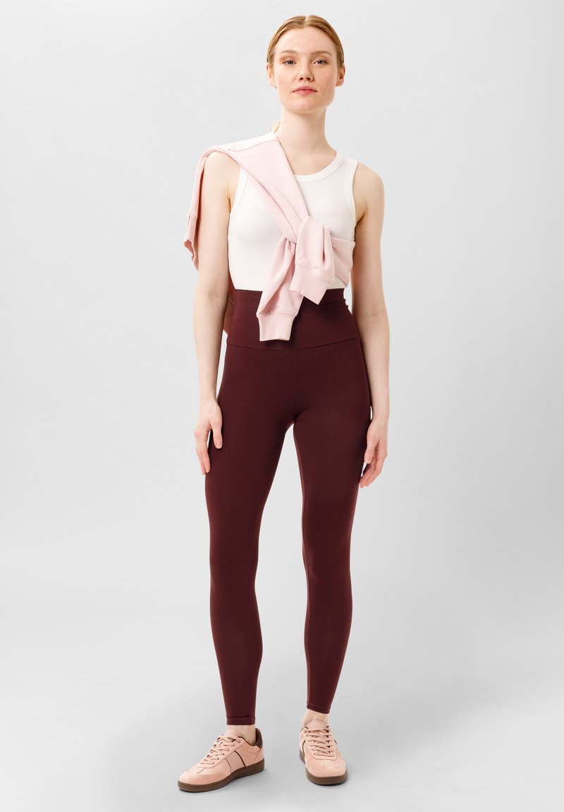 Débardeur blanc, leggings bordeaux taille haute, et hoodie zippé rose drapé sur les épaules ; associé à des baskets rose pâle.