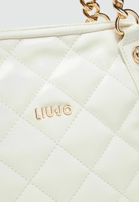 Sac à main en simili cuir crème matelassé avec des détails en métal doré et logo. Il dispose d'une bandoulière en chaîne et d'une texture lisse.