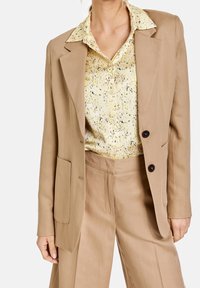 Blazer beige à deux boutons, à revers crantés, avec poches latérales, porté sur une chemise jaune à motifs tachetés, assorti à un pantalon beige.
