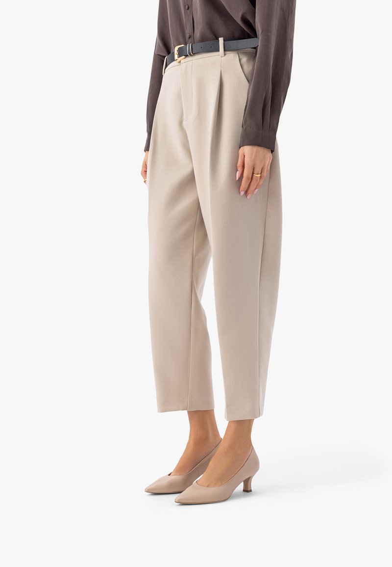 Vrouw die beige op maat gemaakte, enkellange broek draagt, een bruine blouse met lange mouwen, een zwarte riem en beige schoenen met lage hak, staand tegen een witte achtergrond.