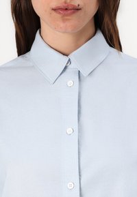 Lyseblå skjorte med krage, laget av glatt stoff, med hvite knapper og pent sydd langs knappestolpen.