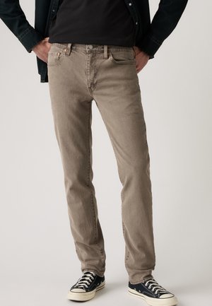 Homme portant un jean droit taupe, des baskets noires aux semelles blanches, une chemise noire et une veste sombre, debout les mains dans les poches.