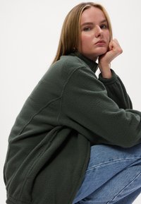 Groene fleece jacket met een hoge kraag en een relaxte pasvorm, met een zachte textuur. Draagt over blauwe spijkerbroeken, wat casual laagjes benadrukt.