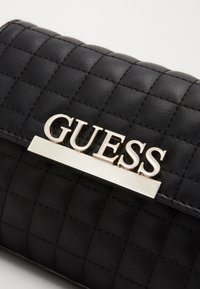 Czarny pikowany portfel z ekoskóry z srebrną plakietką z logo "GUESS" oraz poziomym paskiem dekoracyjnym. Charakteryzuje się fakturowanym wzorem i eleganckim designem.