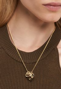 Guldkæde med lagdelte abstrakte guldpendanter båret af en person i en brun ribbet ærmeløs top.