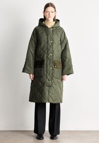 Prošívaný dlouhý khaki zelený péřový kabát s kapucí, zdobený zlatým kováním a dvěma tmavými kapsami. Kombinován s černými kalhotami a botami.
