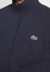 Giacca a zip blu navy con colletto rialzato e piccolo logo di coccodrillo verde sul lato sinistro del petto.
