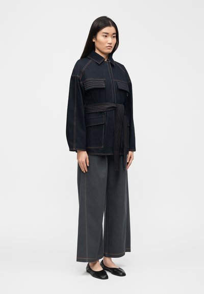 Γυναίκα που φοράει μια σκουρόχρωμη τζιν ζακέτα με ζώνη και μεγάλες τσέπες, καθώς και σκουρόχρωμο παντελόνι με φαρδιά πόδια και αντίθετες ραφές, στέκεται μπροστά σε απλό γκρι φόντο.