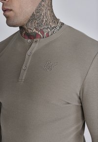 Kačast dugi rukav henley majica s teksturiranim rebrastim uzorkom, prednjim gumbima i malim vezom loga na prsima.