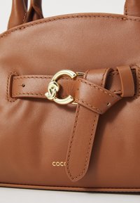 Borsa a mano in pelle marrone con un accento in hardware circolare dorato e un dettaglio a tracolla annodato. Logo in rilievo alla base.