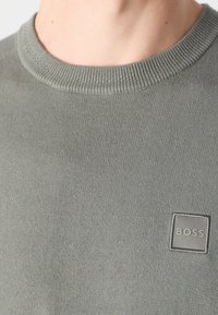 Lähikuva henkilöstä, jolla on päällään harmaa neulepaita, jossa on ribbaus kaula-aukossa ja pieni "BOSS"-appika. 