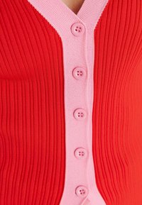 Roter gerippter Cardigan mit pinkem V-Ausschnitt und Knopfleiste, versehen mit sechs pinken Knöpfen entlang der Mitte. Strukturierter Strickstoff.