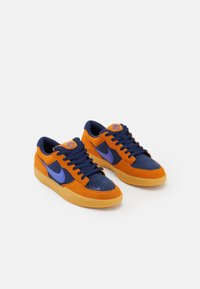 Baskets Nike avec un mélange de daim orange et de mesh bleu foncé. Présentent un swoosh violet et une semelle extérieure en caoutchouc couleur gomme.  Bout perforé pour la respirabilité.