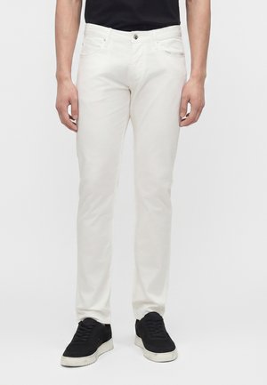 Férfi, aki testhez simuló fehér nadrágot és fekete tornacipőt visel, egy egyszerű világos háttér előtt áll.