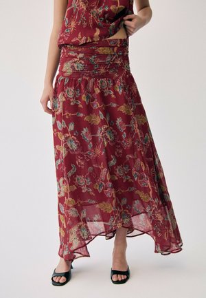 Frau trägt einen roten Maxirock mit Blumenmuster und passendem Oberteil sowie schwarze offene High Heels, steht vor einem einfachen Hintergrund.