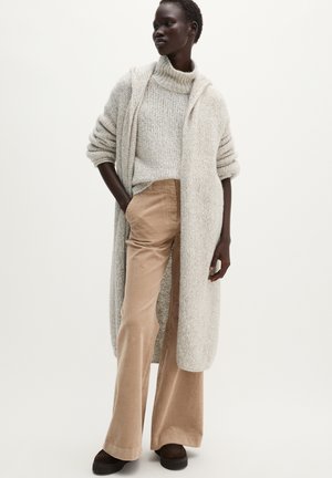 Donna che indossa pantaloni in velluto beige, maglione dolcevita a maglia grossa, cardigan lungo aperto e scarpe scure con plateau, in piedi davanti a uno sfondo semplice.