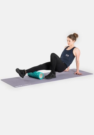 Rolling foam roller, teal with textured spikes, on a gray exercise mat. A person in black leggings and tank top demonstrates use.