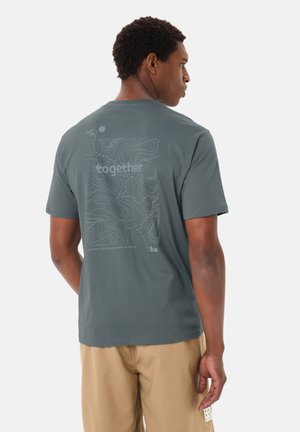 Uomo che indossa una maglietta grigio-verde con design di mappa topografica e la parola "insieme" sul retro, abbinata a pantaloncini beige.